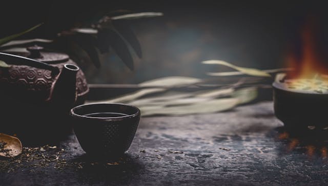 Dark tea background with black iron asian teapot and mug of hot tea on table at dark wall. Copy space for your design. Authentic vintage style. Traditional tea ceremony arrangement | Image Credit: © VICUSCHKA - stock.adobe.com